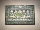A decorative ceramic tile panel depicting a detailed scene of people sitting and interacting, possibly engaged in a banquet or social gathering. The artwork features intricate floral and geometric patterns in the border, with figures adorned in traditional attire, detailed with vivid colors.