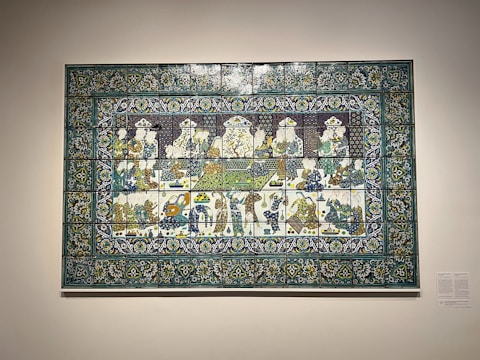 A decorative ceramic tile panel depicting a detailed scene of people sitting and interacting, possibly engaged in a banquet or social gathering. The artwork features intricate floral and geometric patterns in the border, with figures adorned in traditional attire, detailed with vivid colors.