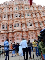An ornate pink sandstone facade features numerous small windows with intricate latticework, resembling the historical architecture typical of Jaipur. A group of people stand in front, some facing the building while others engage in conversation or take photos. The sky is overcast, contributing to a subdued ambiance.