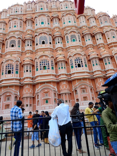 An ornate pink sandstone facade features numerous small windows with intricate latticework, resembling the historical architecture typical of Jaipur. A group of people stand in front, some facing the building while others engage in conversation or take photos. The sky is overcast, contributing to a subdued ambiance.