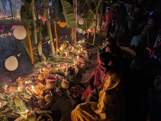 A nighttime scene featuring a traditional ritual setup, with offerings arranged on the ground, surrounded by candles and various food items, possibly fruits and vegetables. People are seated nearby, wearing colorful garments. The area is decorated with banana leaves, marigold garlands, and hanging paper decorations, creating a festive atmosphere.
