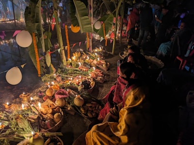 A nighttime scene featuring a traditional ritual setup, with offerings arranged on the ground, surrounded by candles and various food items, possibly fruits and vegetables. People are seated nearby, wearing colorful garments. The area is decorated with banana leaves, marigold garlands, and hanging paper decorations, creating a festive atmosphere.