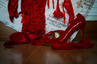 A collection of vibrant red lingerie and high-heeled shoes, with delicate lace details, is arranged on a wooden surface. The background features a fabric with text and butterfly illustrations, enhancing the romantic and elegant theme.