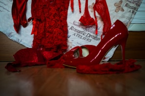 A collection of vibrant red lingerie and high-heeled shoes, with delicate lace details, is arranged on a wooden surface. The background features a fabric with text and butterfly illustrations, enhancing the romantic and elegant theme.