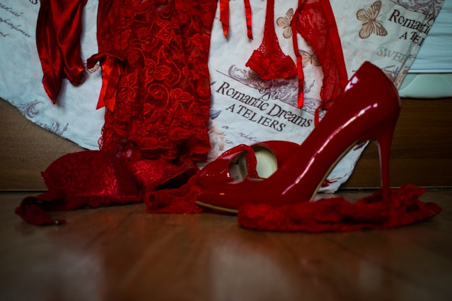 A collection of vibrant red lingerie and high-heeled shoes, with delicate lace details, is arranged on a wooden surface. The background features a fabric with text and butterfly illustrations, enhancing the romantic and elegant theme.
