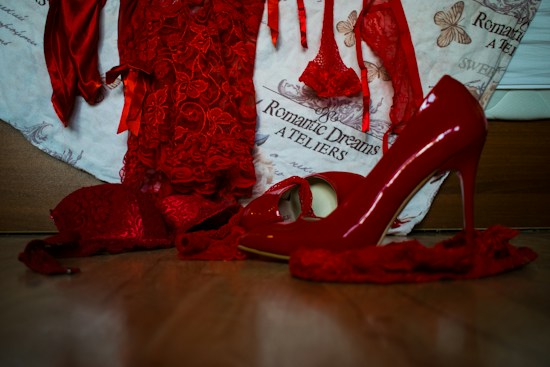 A collection of vibrant red lingerie and high-heeled shoes, with delicate lace details, is arranged on a wooden surface. The background features a fabric with text and butterfly illustrations, enhancing the romantic and elegant theme.