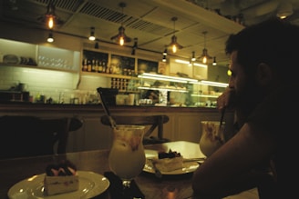 A dimly lit café interior featuring a person sitting at a table with two desserts and drinks. The background showcases a counter with various bottles, a display case, and hanging industrial-style lights.