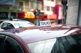 A red car is visible with a white and blue taxi sign on its roof. In the background, there are blurred images of buildings and another vehicle, indicating an urban setting.