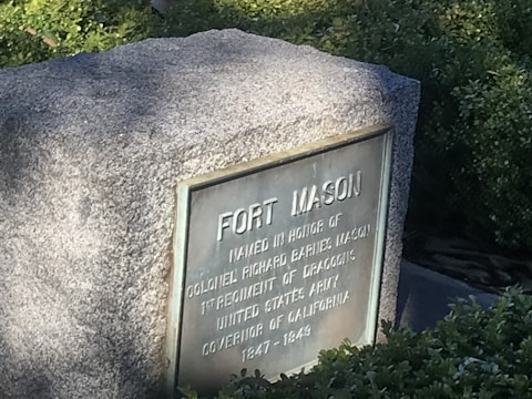 A stone monument with an engraved metal plaque that reads 'FORT MASON' and honors Colonel Richard Barnes Mason. The monument is surrounded by greenery and bathed in sunlight, casting shadows on its surface.