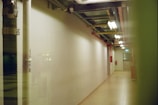 A dimly lit corridor with overhead lighting and exposed piping runs along the ceiling. The walls are plain and the floor is tiled. A fire extinguisher is mounted on the right wall, and the end of the hallway has a closed door with a small window.
