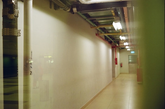 A dimly lit corridor with overhead lighting and exposed piping runs along the ceiling. The walls are plain and the floor is tiled. A fire extinguisher is mounted on the right wall, and the end of the hallway has a closed door with a small window.