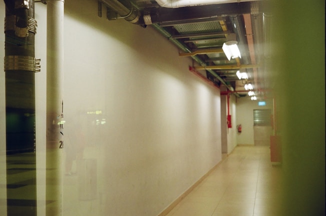 A dimly lit corridor with overhead lighting and exposed piping runs along the ceiling. The walls are plain and the floor is tiled. A fire extinguisher is mounted on the right wall, and the end of the hallway has a closed door with a small window.