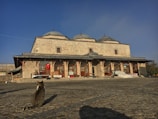 A stone building with three domes, featuring arched windows and a Turkish flag hanging from one facade. A cobblestone courtyard stretches in front of the building, with a tabby cat sitting in the foreground gazing towards the building.
