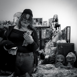 A person with curly hair and glasses, wearing casual clothing and a beanie, stands among shelves of toys and dolls. They are holding a plastic-wrapped object and appear to be shopping in a cluttered toy store.