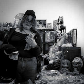 A person with curly hair and glasses, wearing casual clothing and a beanie, stands among shelves of toys and dolls. They are holding a plastic-wrapped object and appear to be shopping in a cluttered toy store.