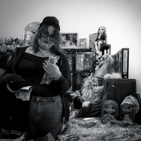 A person with curly hair and glasses, wearing casual clothing and a beanie, stands among shelves of toys and dolls. They are holding a plastic-wrapped object and appear to be shopping in a cluttered toy store.