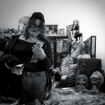 A person with curly hair and glasses, wearing casual clothing and a beanie, stands among shelves of toys and dolls. They are holding a plastic-wrapped object and appear to be shopping in a cluttered toy store.