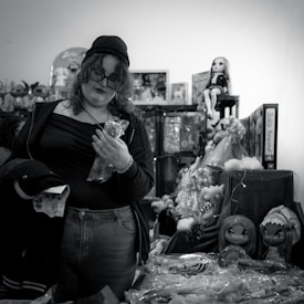 A person with curly hair and glasses, wearing casual clothing and a beanie, stands among shelves of toys and dolls. They are holding a plastic-wrapped object and appear to be shopping in a cluttered toy store.