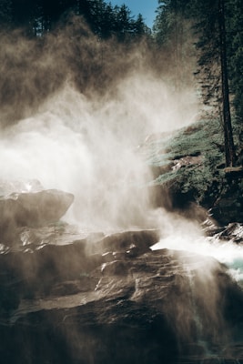 A misty waterfall cascading down a rocky terrain surrounded by dense forest. Sunlight filters through creating a mystical atmosphere.