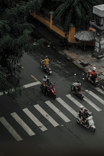 Motorcyclists navigate a wet, urban street, crossing a zebra crossing. The scene is overseen by lush green foliage and a yellow wall with decorative ironwork. A person holds an umbrella, suggesting ongoing or recent rain. Several scooters are in motion, with riders wearing helmets.