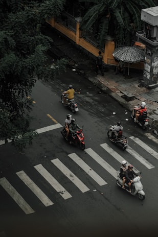 Motorcyclists navigate a wet, urban street, crossing a zebra crossing. The scene is overseen by lush green foliage and a yellow wall with decorative ironwork. A person holds an umbrella, suggesting ongoing or recent rain. Several scooters are in motion, with riders wearing helmets.