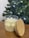 A glass jar candle with a wooden lid rests on a wooden surface. The candle wax is white and has a decorative design on the surface. In the background, there is a Christmas tree adorned with lights, creating a festive atmosphere.
