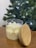 A glass jar candle with a wooden lid rests on a wooden surface. The candle wax is white and has a decorative design on the surface. In the background, there is a Christmas tree adorned with lights, creating a festive atmosphere.