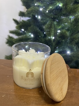 A glass jar candle with a wooden lid rests on a wooden surface. The candle wax is white and has a decorative design on the surface. In the background, there is a Christmas tree adorned with lights, creating a festive atmosphere.