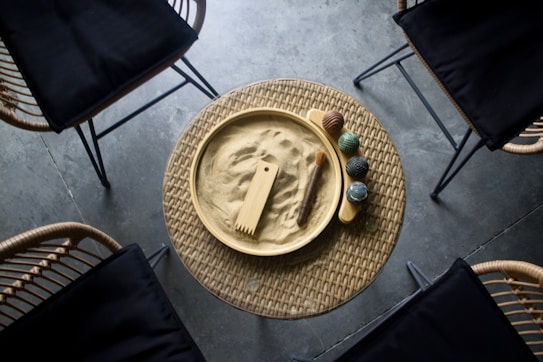 A circular wicker table holds a small zen garden with raked sand, a wooden rake, and a brush. Several textured spheres sit beside the garden. Surrounding the table are four black-cushioned chairs on a concrete floor.