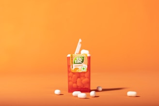 A vibrant orange backdrop highlights a partially opened pack of Tic Tac mints. The pack is labeled with the flavor 'Orange', and several white mints are scattered around the container.