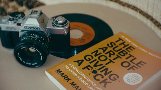 An analog camera, a vinyl record, and a book titled 'The Subtle Art of Not Giving a F*ck' are placed on a flat surface, suggesting a blend of photography, music, and literature.