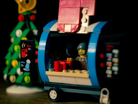 A small Lego scene features a figurine inside a colorful kiosk or food truck with bright elements made of blocks. The structure has blue and pink hues, and there is a holiday-themed Lego tree in the background. The figurine appears to be a vendor, smiling and surrounded by miniature cups and items. An illustrated sign on the side shows images of beverages.