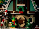 A Lego scene featuring a holiday setting with a small building adorned with a wreath. An elf figurine dressed in green with a red scarf stands in front. A signpost next to the elf indicates directions to the North Pole and Workshop. The scene includes festive elements like colored lights and a Christmas tree.