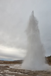 A geyser erupts forcefully, sending a towering column of water and steam into the cloudy sky. The surrounding landscape is flat with some distant hills and sparse vegetation.