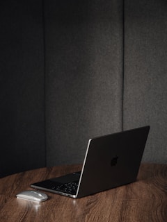 A sleek, closed laptop with an Apple logo sits on a wooden table alongside a wireless mouse. The background consists of dark upholstered panels, creating a minimalist and modern setting.