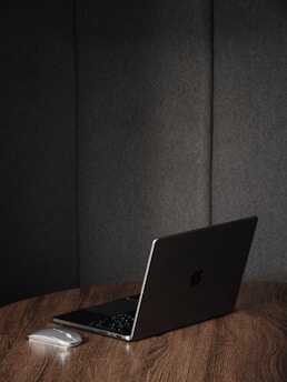 A sleek, closed laptop with an Apple logo sits on a wooden table alongside a wireless mouse. The background consists of dark upholstered panels, creating a minimalist and modern setting.