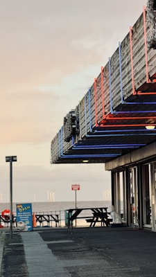 An outdoor seating area near a waterfront, featuring picnic tables and a vintage-looking cafe or restaurant with neon lights in blue and red. The sky appears overcast, adding a subdued ambiance. There's a sign advertising hot drinks and snacks, and wind turbines are visible in the distance across the water.