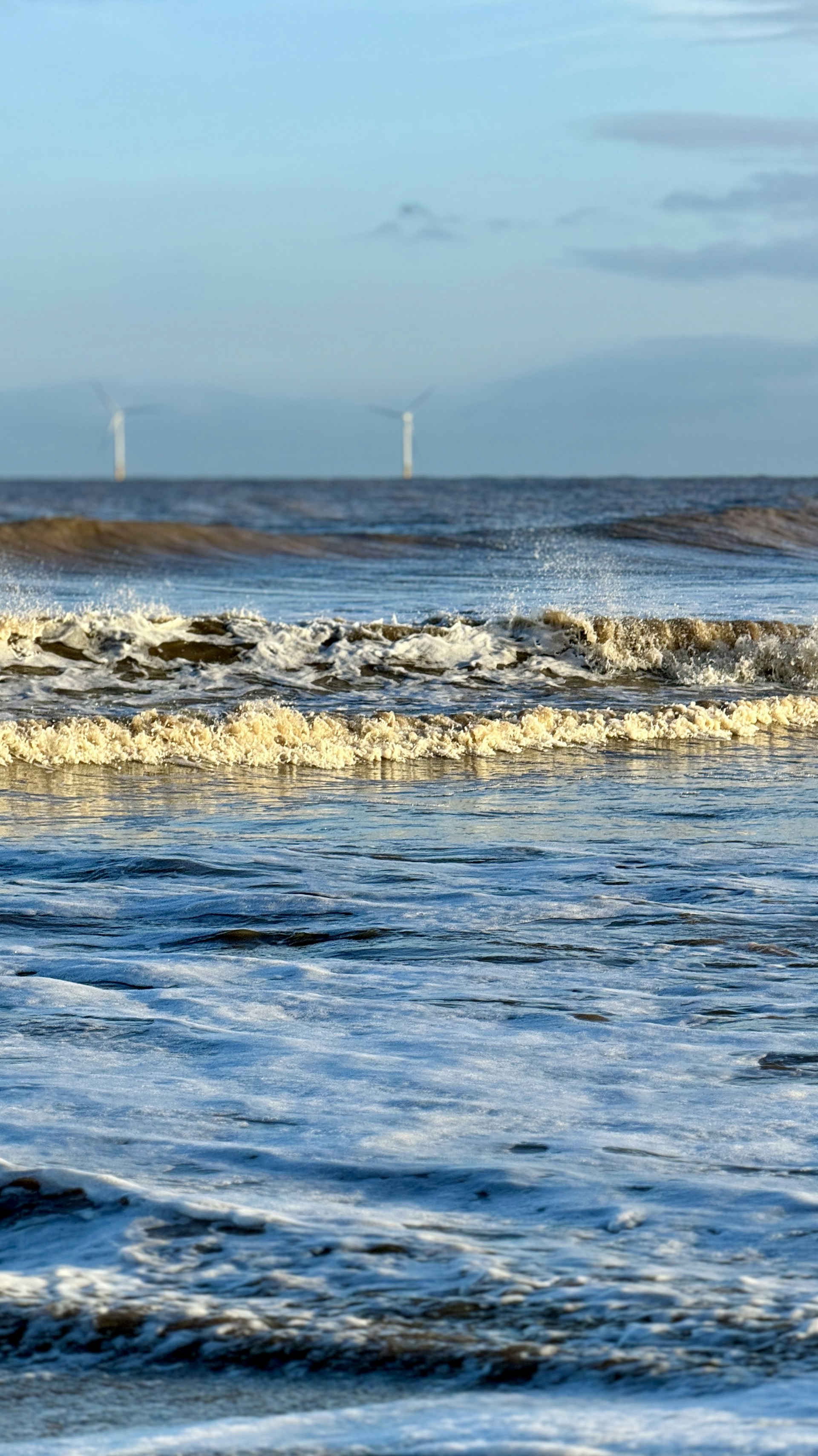 Waves gently crash onto the shore, reflecting the soft blues and whites of the ocean. Wind turbines are visible in the distance against a pale sky, suggesting a blend of natural beauty and sustainable technology.