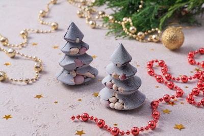 Two cone-shaped, layered pastries resembling Christmas trees are decorated with colorful nuts or candies. They're surrounded by festive gold and red bead garlands and small golden star confetti on a textured surface. A sprig of greenery and a golden ornament add a holiday touch.