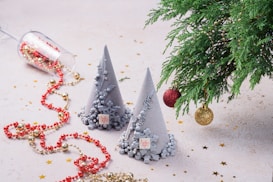 Two cone-shaped decorations with intricate designs are positioned prominently, resting on a light surface sprinkled with gold stars. Nearby, a tipped glass tumbler spills out a strand of red and gold beads. On the right, a lush green branch displays red and gold baubles. The overall arrangement suggests a festive, celebratory setting.