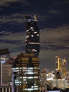 A cityscape at night featuring a modern skyline with skyscrapers illuminated by artificial lights. The tallest building has a distinctive, pixelated architecture with irregularly stacked segments. The sky is partly cloudy, creating a dramatic backdrop.