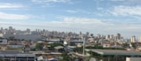 A cityscape featuring a mix of residential and commercial buildings under a partly cloudy sky. The foreground includes some industrial or warehouse structures with tree-lined streets and parked cars. In the background, modern high-rise buildings dominate the skyline.
