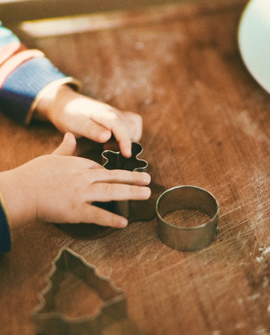 Small hands working with cookie cutters on a wooden surface, including a gingerbread man and a tree shape.