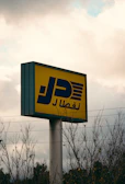 A tall signpost with a rectangular yellow sign featuring bold blue Arabic script and graphic elements is visible against a cloudy sky. Sparse leafless branches partially obscure the base of the sign. Power lines stretch horizontally across the scene, adding an industrial feel.