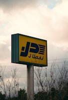 A tall signpost with a rectangular yellow sign featuring bold blue Arabic script and graphic elements is visible against a cloudy sky. Sparse leafless branches partially obscure the base of the sign. Power lines stretch horizontally across the scene, adding an industrial feel.
