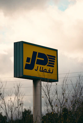 A tall signpost with a rectangular yellow sign featuring bold blue Arabic script and graphic elements is visible against a cloudy sky. Sparse leafless branches partially obscure the base of the sign. Power lines stretch horizontally across the scene, adding an industrial feel.