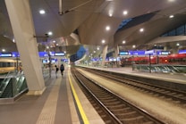 A modern train station platform with sleek architectural design. Overhead lighting illuminates the area with a bright, clean appearance. There are multiple train tracks visible, and people are walking along the platform. Digital signs and escalators are present, indicating a large station environment. Trains are stationed in the background, colored predominantly in red and yellow.