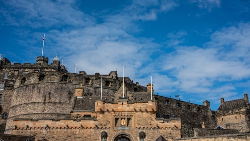 A historic stone castle with towering walls and battlements is set against a bright blue sky. The structure is accented by various flags flying atop its towers and features a central entrance adorned with heraldic symbols.