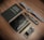 A neatly arranged set of everyday carry items placed on a wooden surface, consisting of a Field Notes notebook, a wristwatch with a gray strap, a sleek black wallet, a black metal pen, a compact utility knife, and a multi-tool with multiple foldable components.