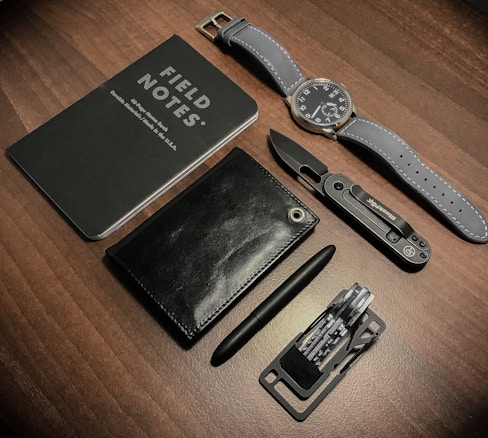 A neatly arranged set of everyday carry items placed on a wooden surface, consisting of a Field Notes notebook, a wristwatch with a gray strap, a sleek black wallet, a black metal pen, a compact utility knife, and a multi-tool with multiple foldable components.